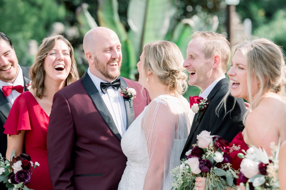 Wedding party members share joyful laughter during outdoor photography session in garden setting.
