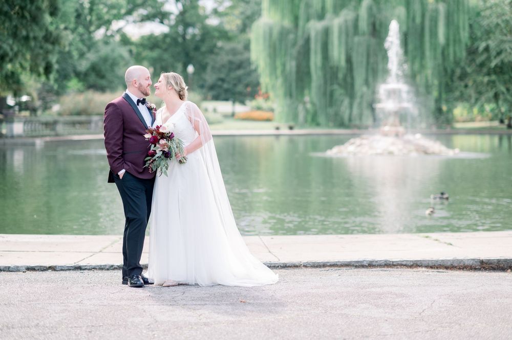 A couple poses for romantic wedding photos near a serene lake with a fountain and weeping willow trees.