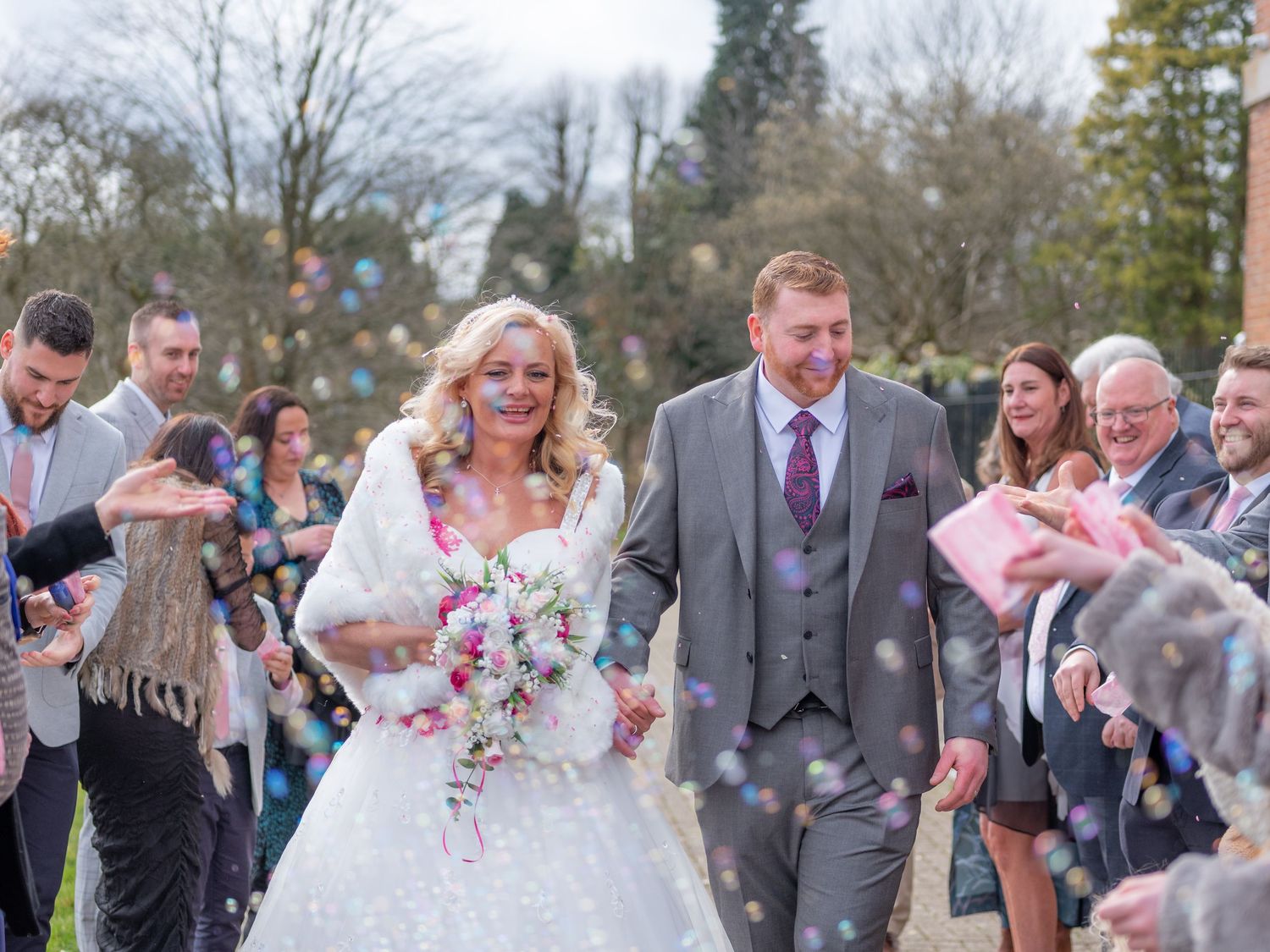 A bride holding a pink and white rose bouquet poses with her groom in a navy suit during an outdoor wedding.