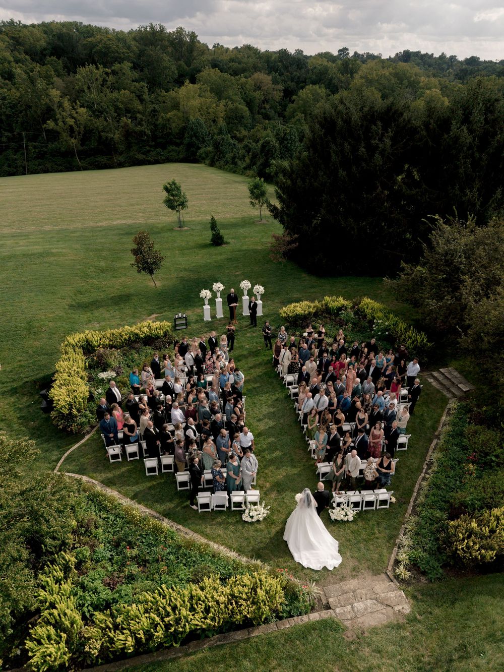 Aerial drone view of an outdoor wedding ceremony with guests seated in a garden surrounded by lush green trees and rolling hills.