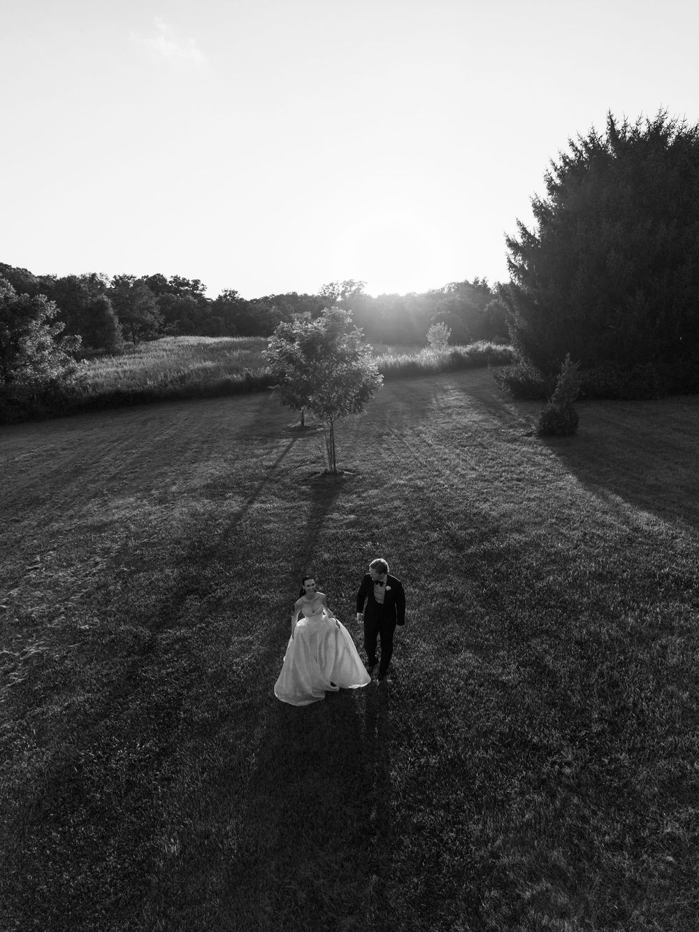 Aerial black and white photo of a bride and groom walking hand in hand through a sunlit green field at sunset.