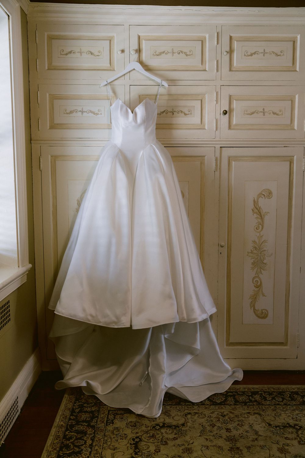 Elegant white strapless ball gown wedding dress hanging on a hanger against a vintage cream armoire.