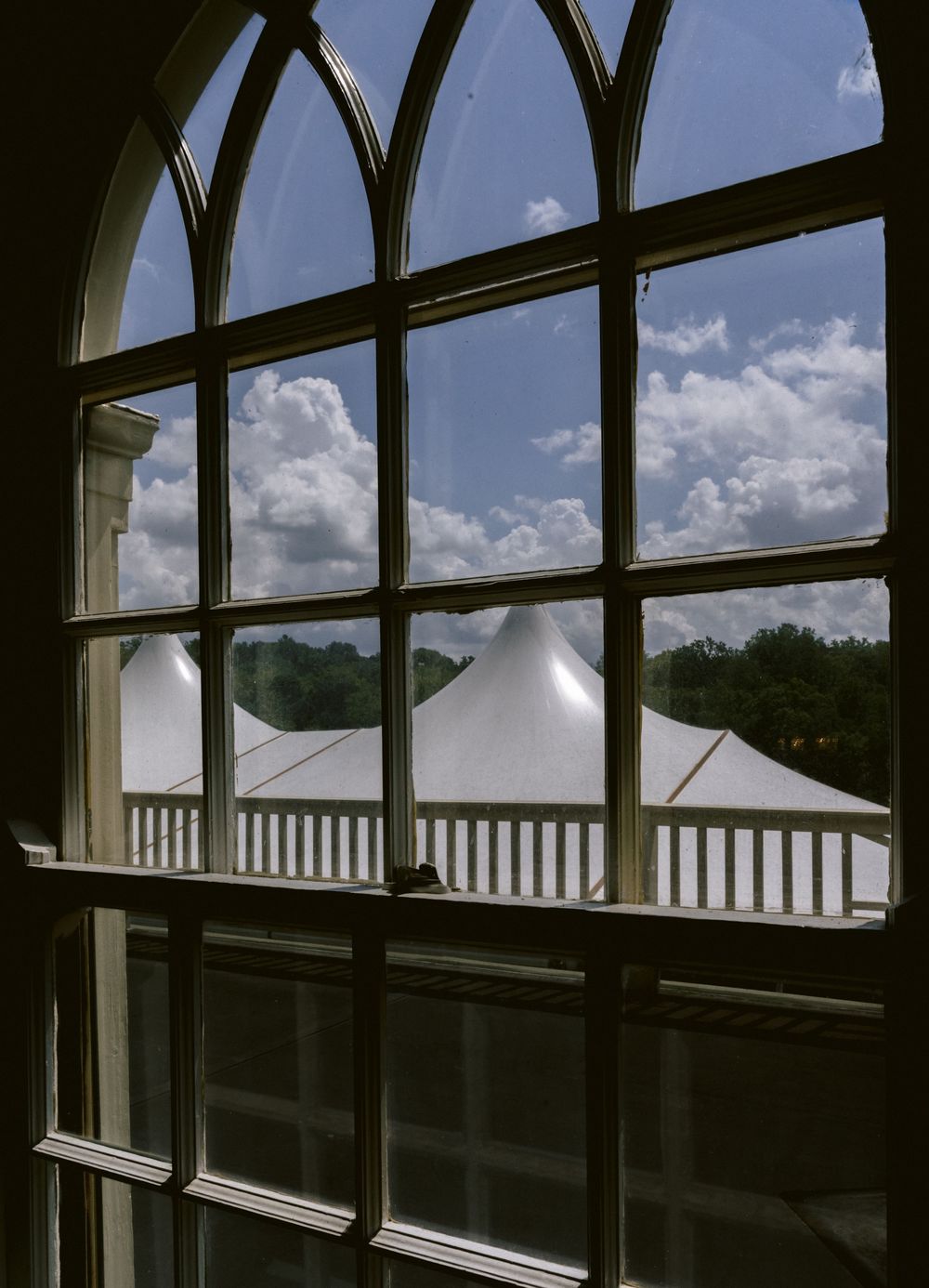 View through arched window of white tent structures under blue sky with clouds, framed by wooden mullions.