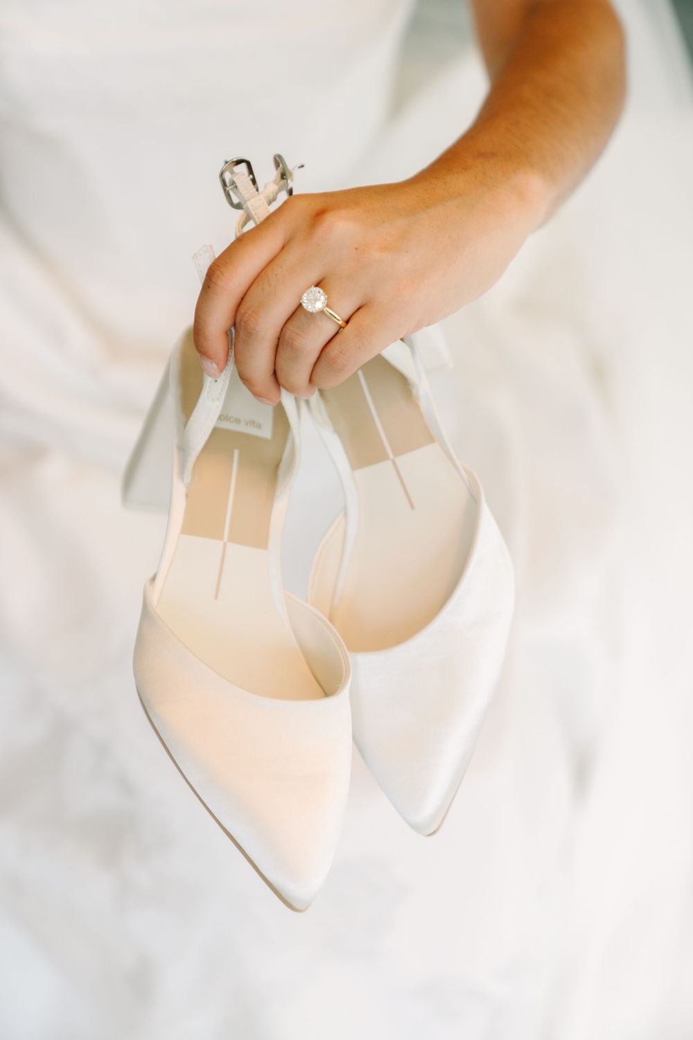 Bride holding white pointed-toe heels with ankle straps, wearing a diamond engagement ring, against a white wedding dress.