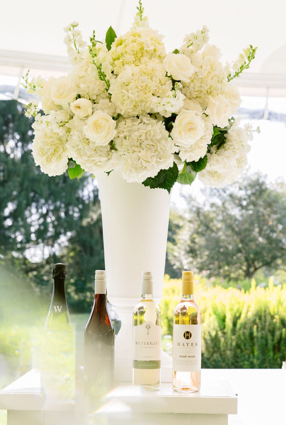 White hydrangeas and roses in tall vase with wine bottles at elegant wedding reception.