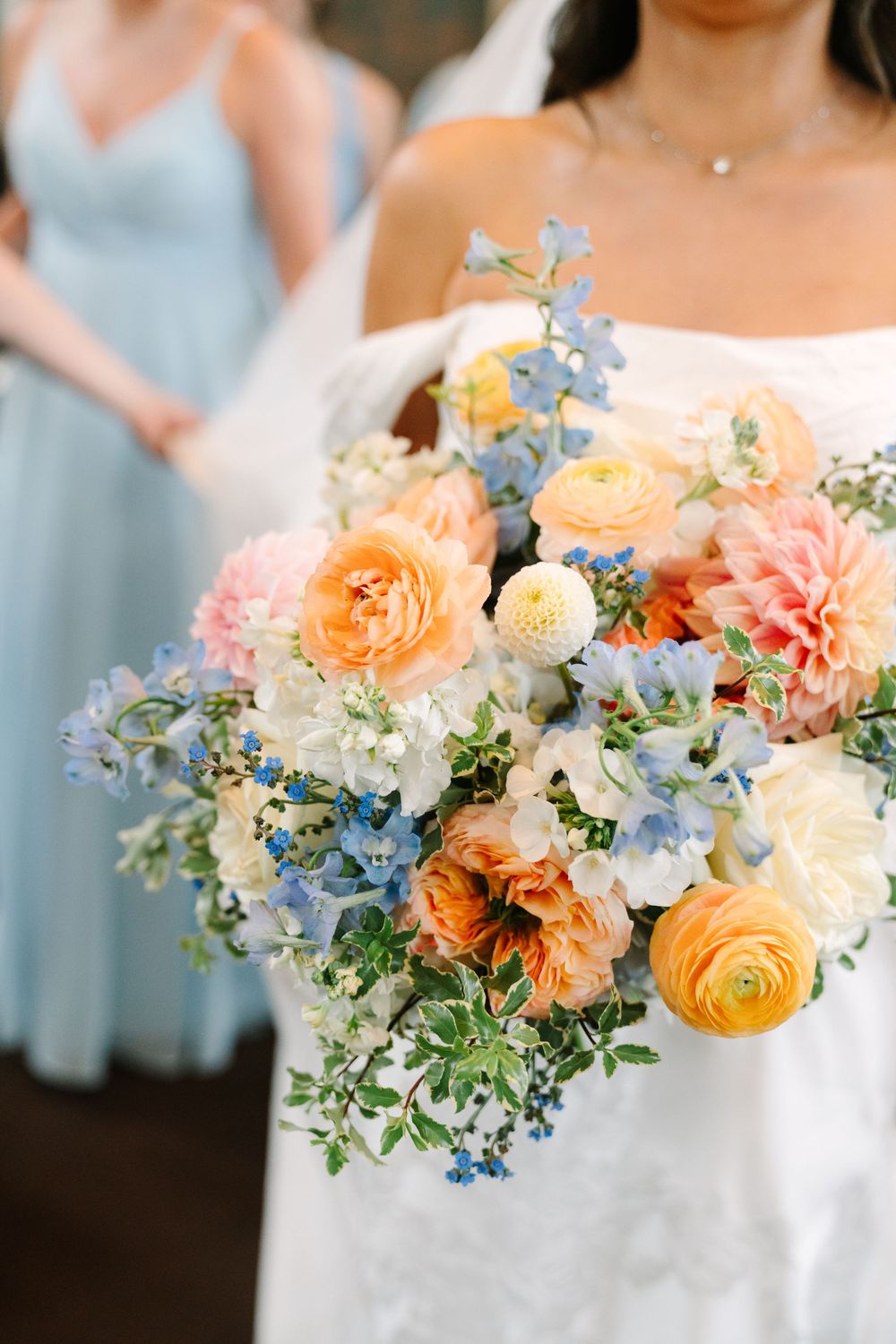 Bride holding a lush wedding bouquet with peach, pink, blue, and white flowers including ranunculus and dahlias.