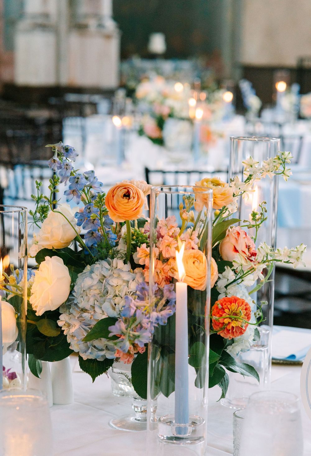 Elegant wedding centerpiece with peach ranunculus, blue hydrangeas, and candles in glass cylinders on a white tablecloth.