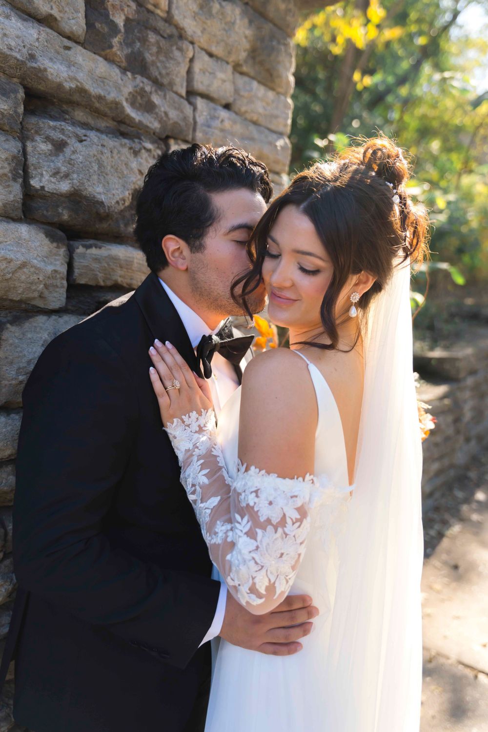 Romantic wedding portrait against stone wall with sunlit natural backdrop.