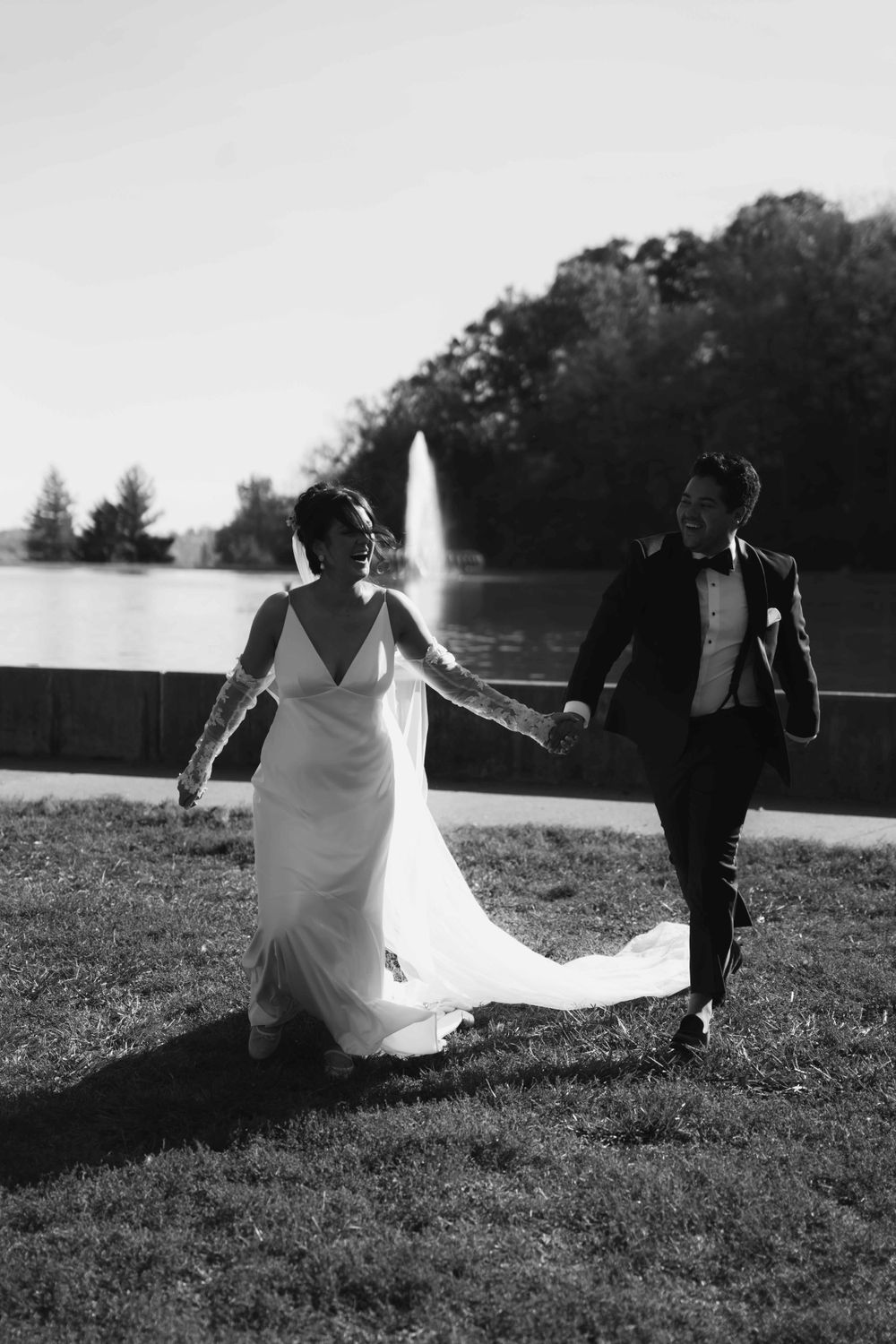 A wedding couple walks hand-in-hand across a lawn with a fountain in the background, captured in black and white.