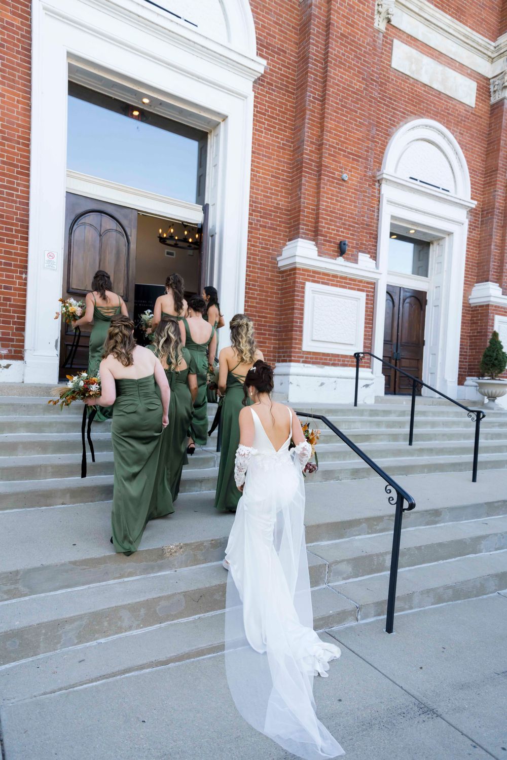 Wedding party entering a red brick church with white trim as bridesmaids in green dresses climb the steps.