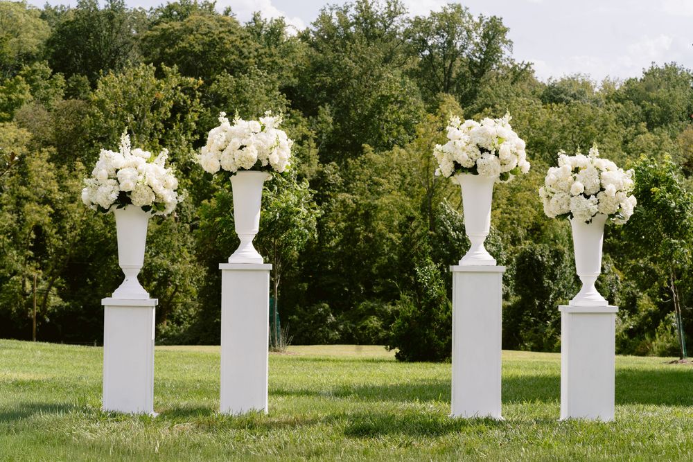 Four white pedestal urns with white floral arrangements on green lawn with trees backdrop for outdoor wedding ceremony.