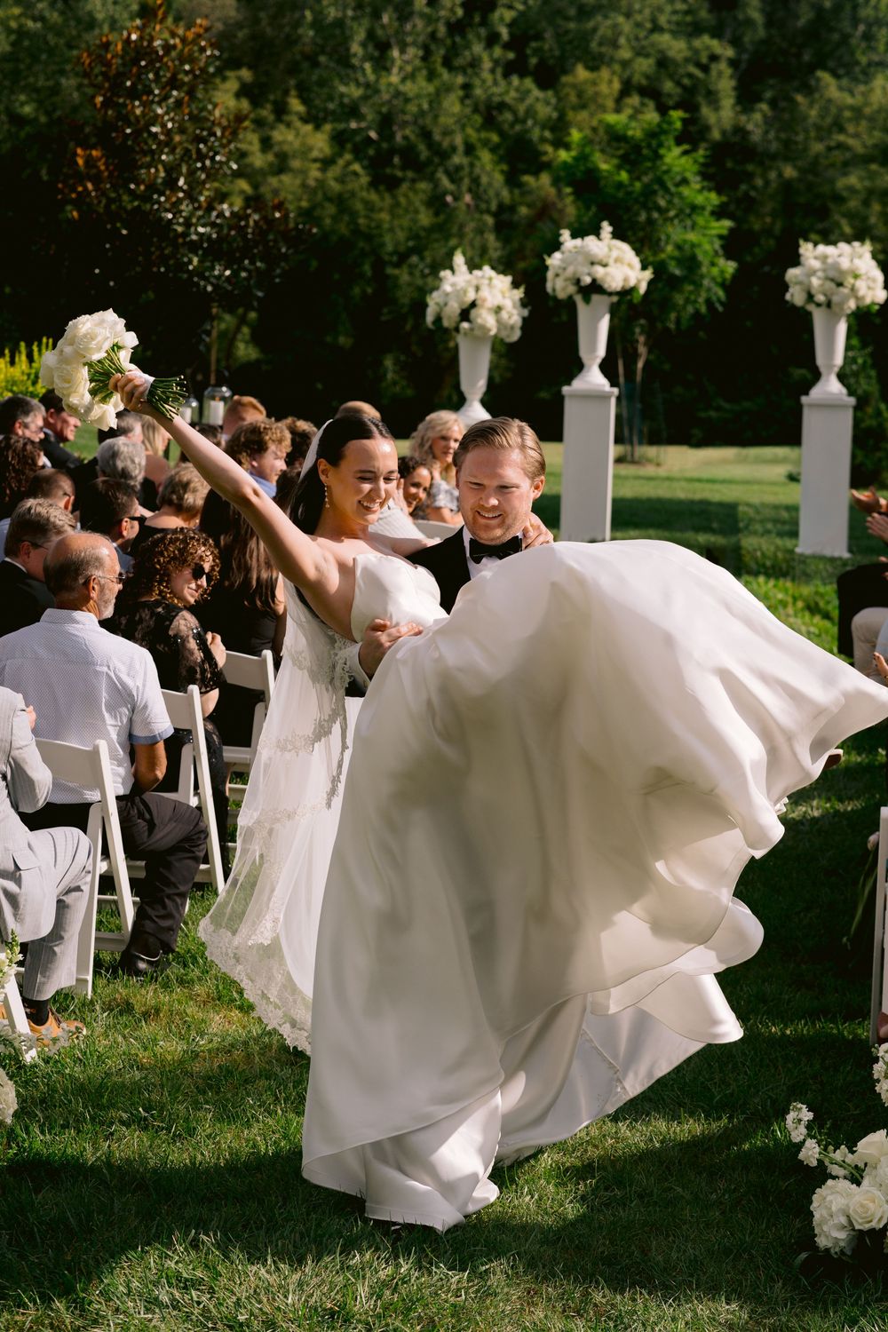 Joyful bride in flowing white gown raises bouquet while groom embraces her during outdoor wedding ceremony recessional.