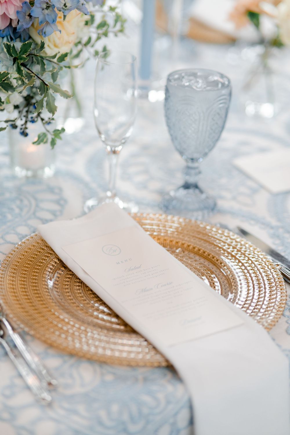 Elegant wedding table setting with gold charger plate, white menu card, blue goblet, and floral centerpiece on blue tablecloth.