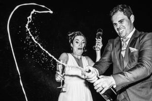 Zwart-witfoto van een bruidspaar dat een champagnefles opent, met een spectaculaire spuitende straal en hilarische gezichtsuitdrukkingen