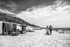 Zwart-witfoto van een bruidspaar dat hand in hand over een rustig strand wandelt, langs strandhuisjes en ontspannen zonnebaders
