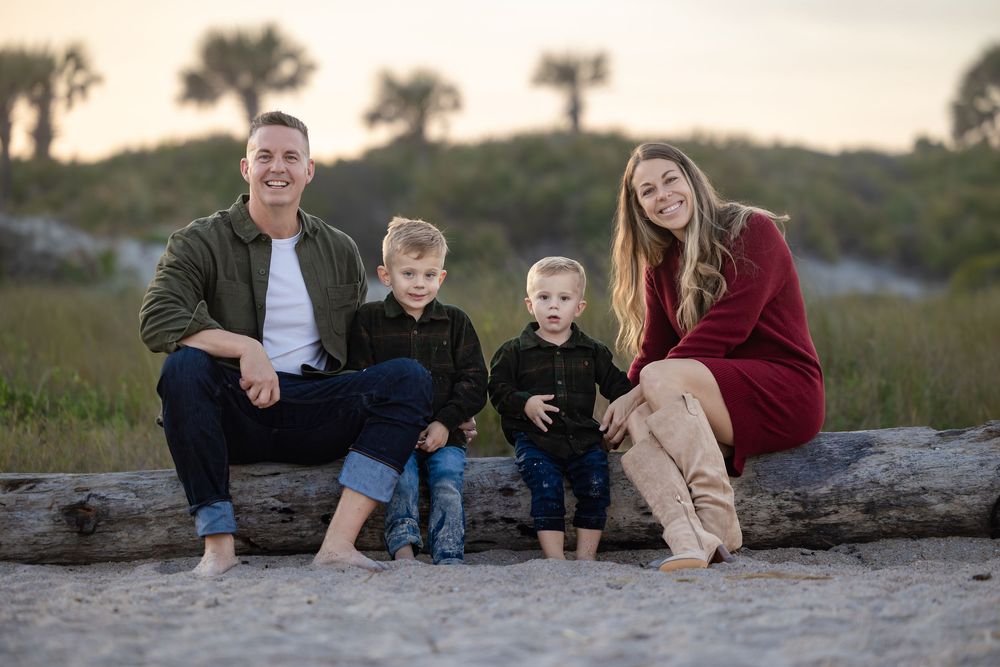 Family relaxing together on driftwood at coastal beach during sunset.