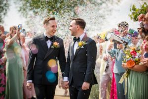 Two grooms in black tuxedos hold hands smiling as colorful confetti falls during their outdoor wedding celebration.