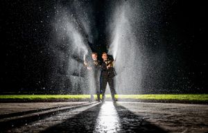 Two firefighters stand boldly in a dramatic water spray at night, illuminated by bright light on a wet surface.
