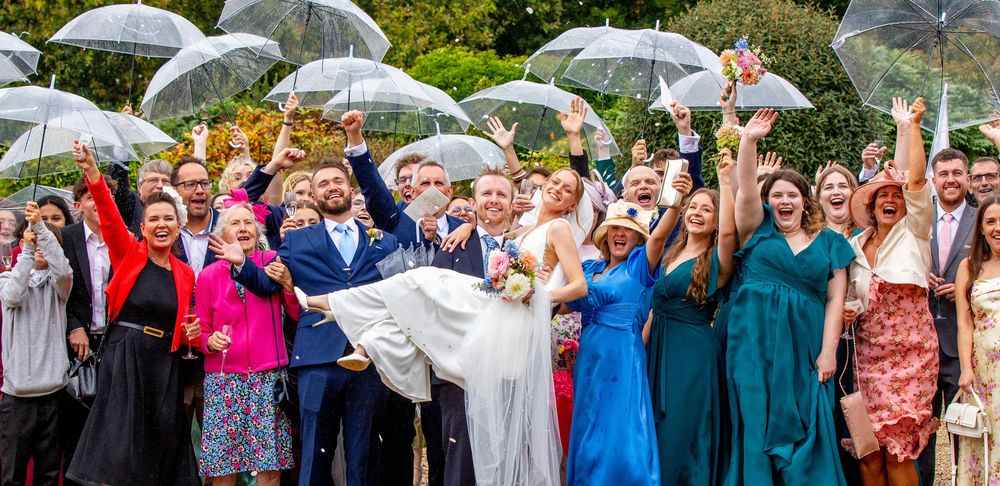 Joyful wedding party celebrating in the rain with clear umbrellas, bride being carried by groom outdoors.