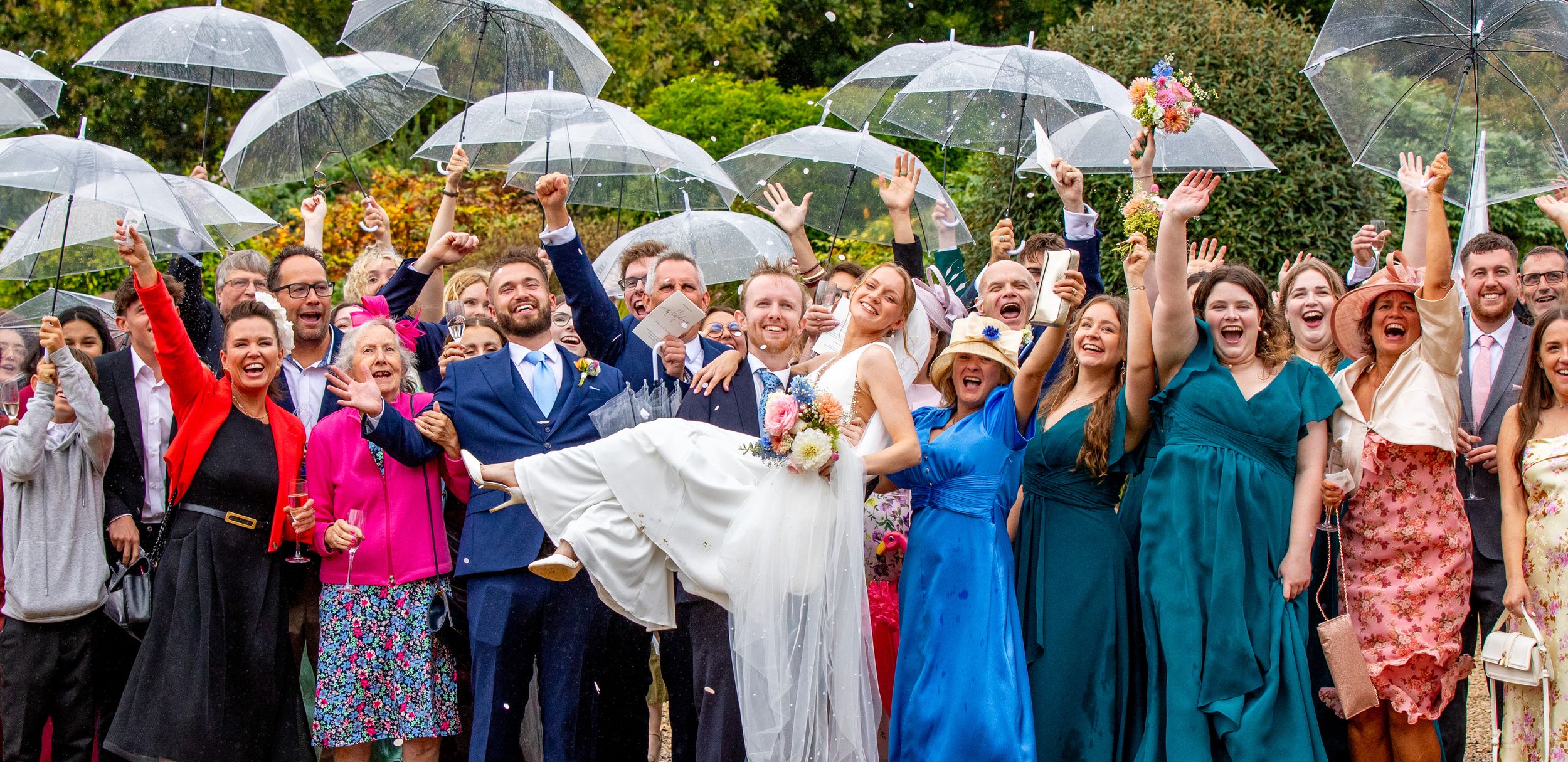 Joyful wedding party celebrating in the rain with clear umbrellas, bride being carried by groom outdoors.