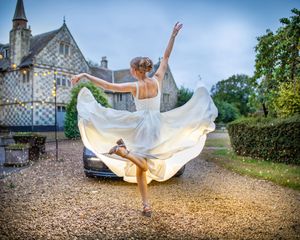 Bride in flowing white gown dancing gracefully outside a historic manor on a gravel driveway.