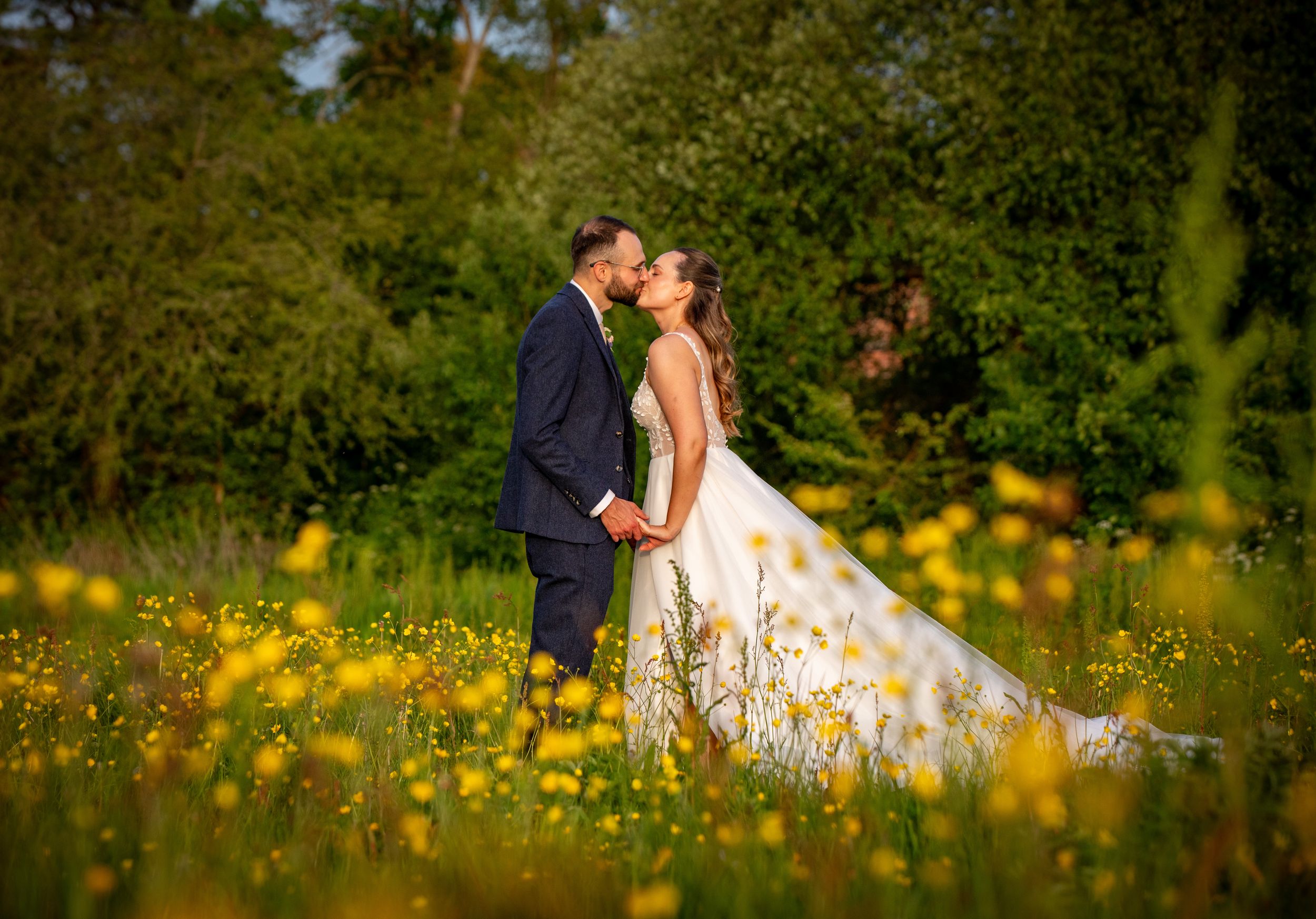 Bride and groom kissing in a sunlit meadow filled with yellow wildflowers, surrounded by lush green trees.