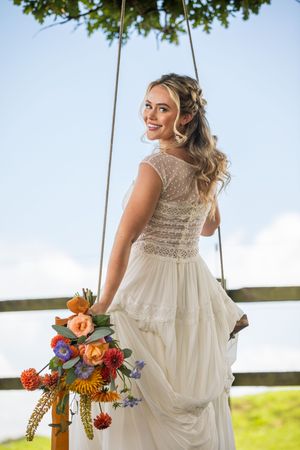 Bride in lace wedding dress sits on rustic wooden swing adorned with vibrant wildflowers on sunny day.