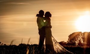 Romantic couple silhouetted against golden sunset sky, embracing in a field, bride in flowing gown with train.