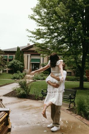 A romantic embrace and lift on a garden pathway outside a stone building with green trees.