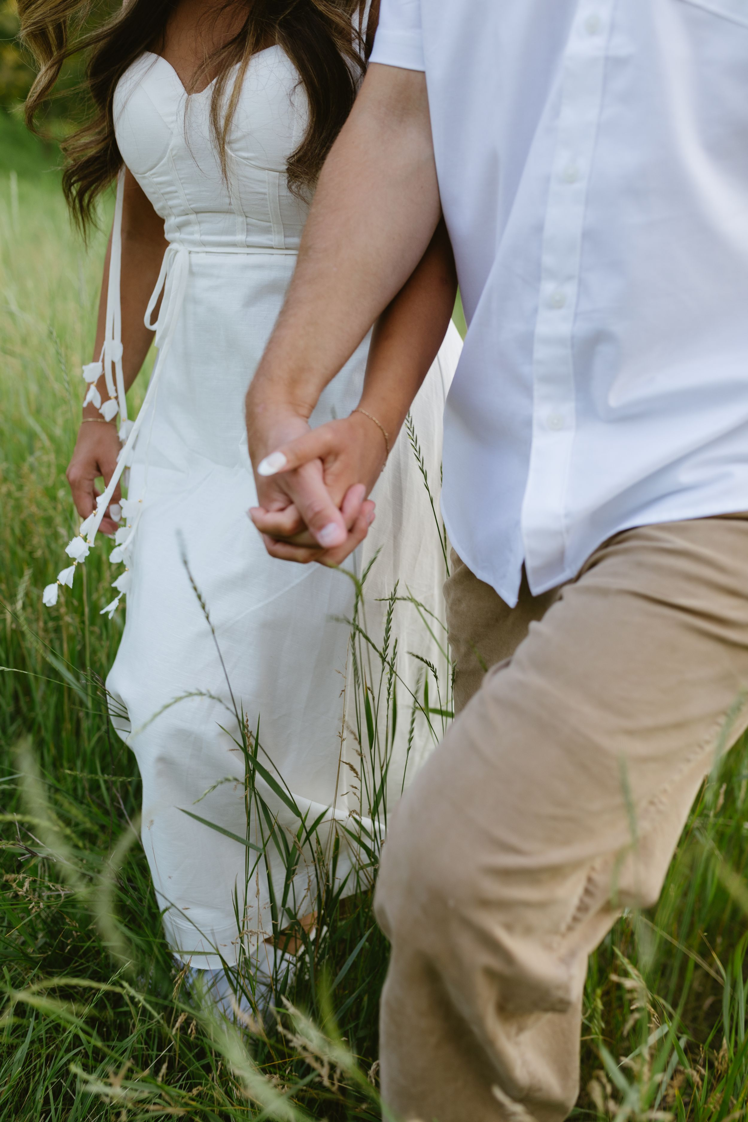 A couple holds hands while walking through tall grass, with focus on white dress and khaki pants.