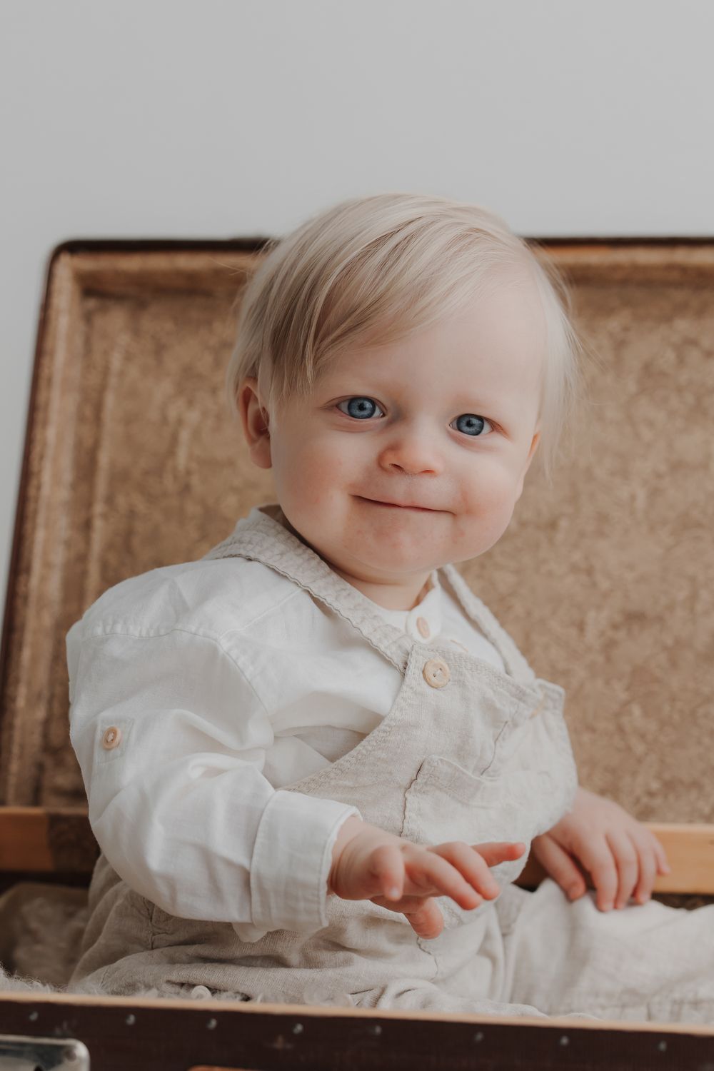 Adorable blue-eyed baby with blonde hair sitting in a vintage suitcase, wearing a white button-up shirt.