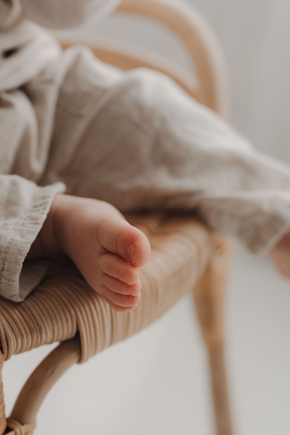 Close-up of tiny newborn baby feet resting on a rattan chair with soft neutral linen fabric.