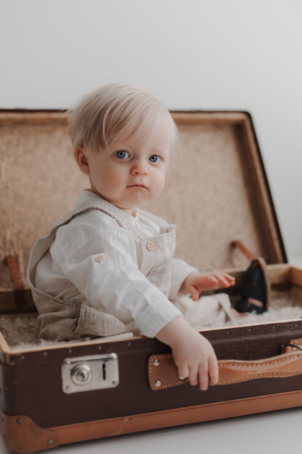 A blue-eyed baby boy in a white shirt sits inside a vintage brown suitcase against a white background.