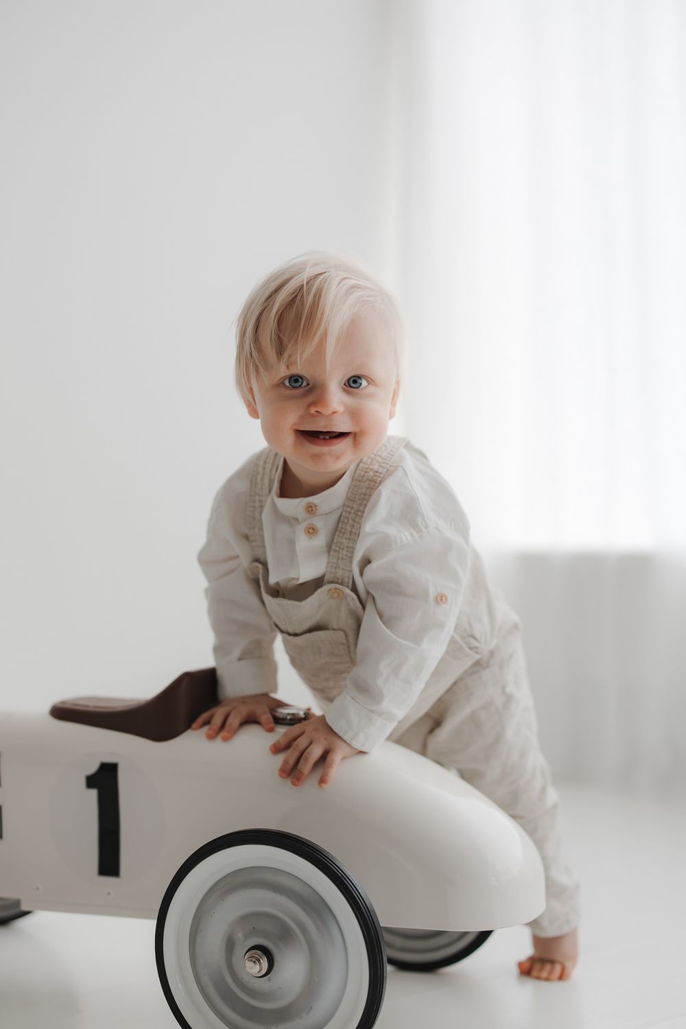 Smiling blonde toddler in beige overalls playing on a white vintage toy race car in a bright white room.