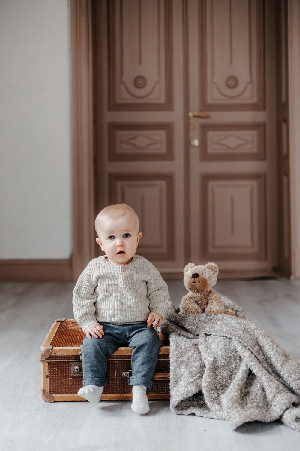 Small child sits on vintage wooden trunk with teddy bear and blanket against brown door in minimalist setting.