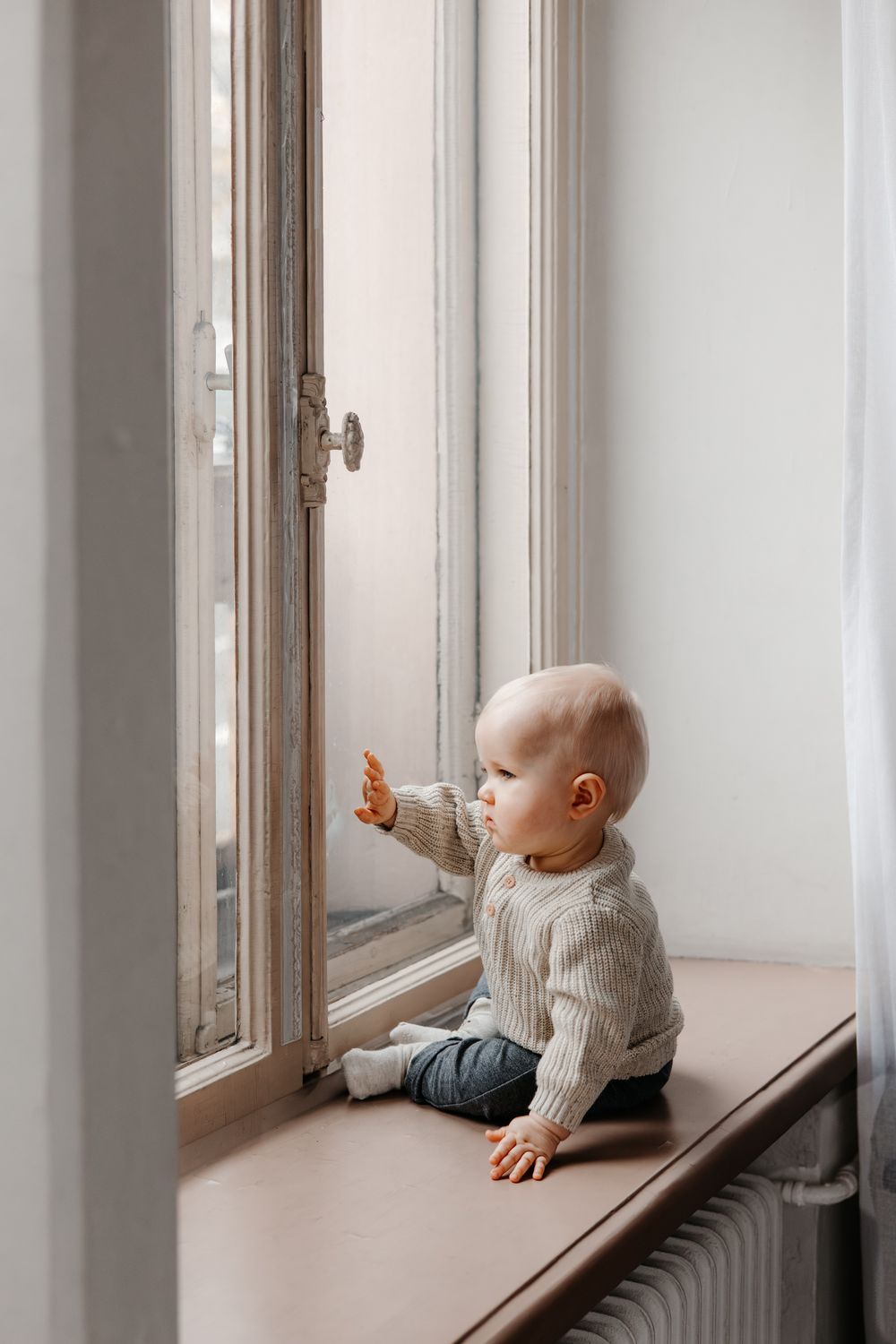Baby in gray sweater gazes out window while sitting on white windowsill in bright interior space.