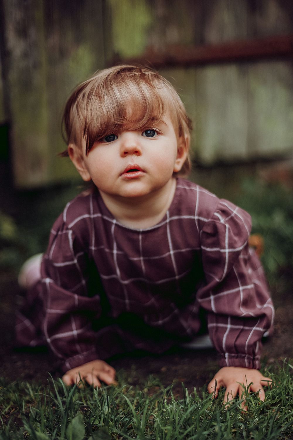 Baby in plaid shirt and overalls crawls on grass near rustic wooden wall.