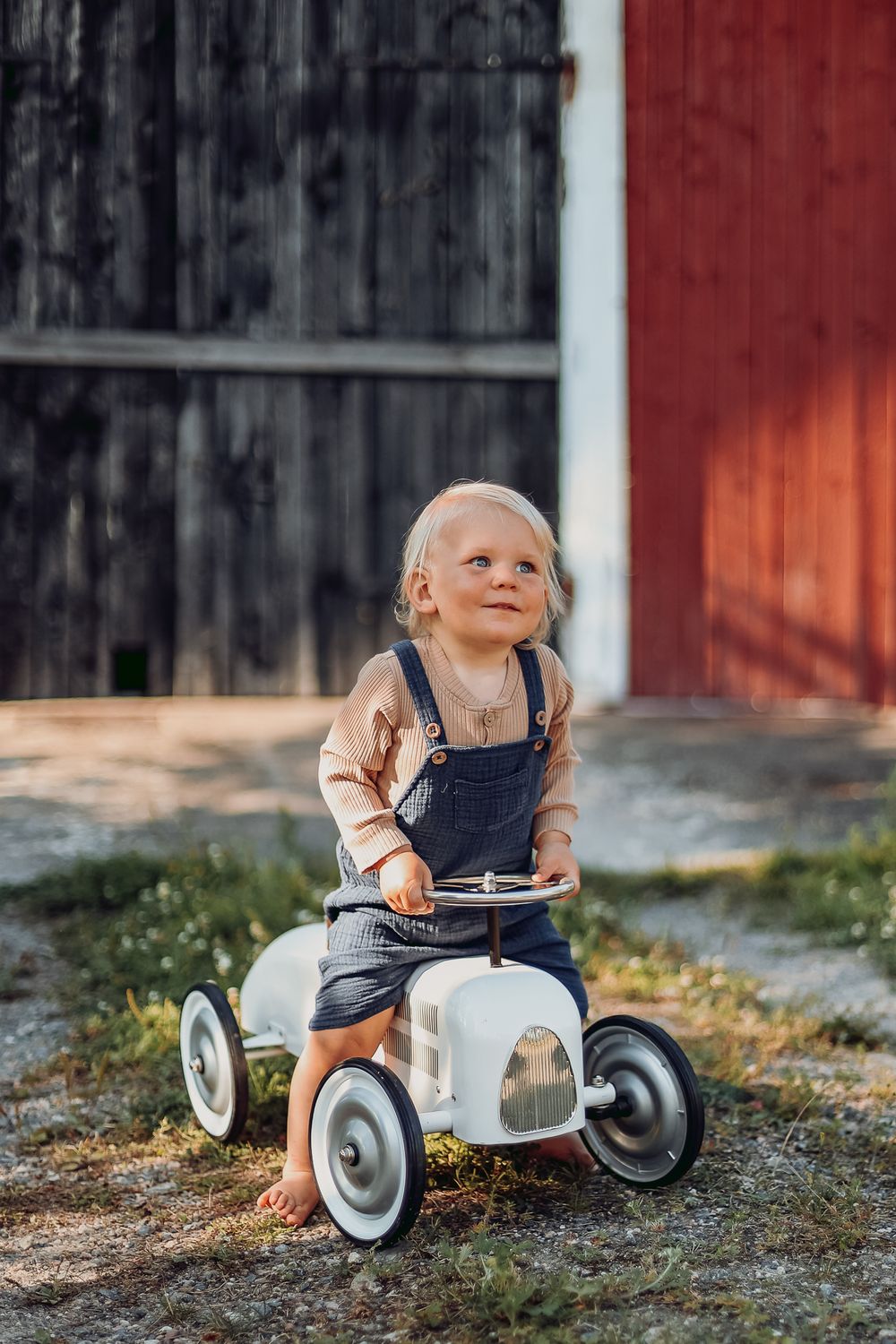 Small child on retro pedal car smiles in front of red barn.