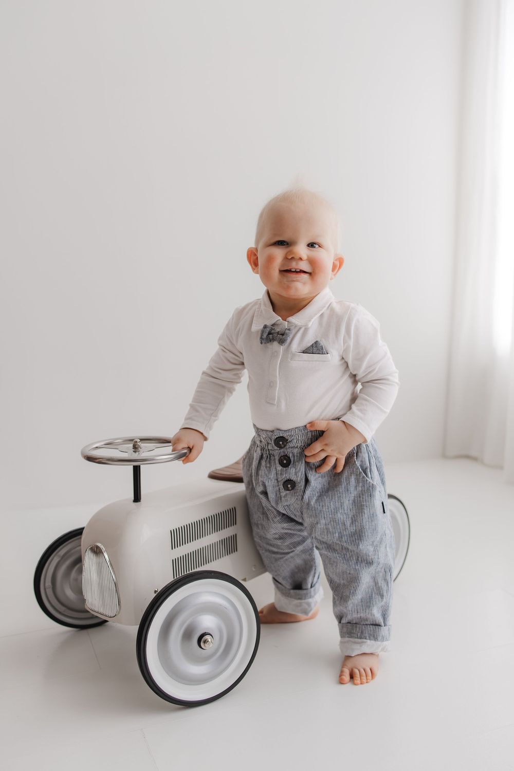 Smiling baby boy in white shirt, bow tie, and gray suspender pants standing next to a vintage white toy car.