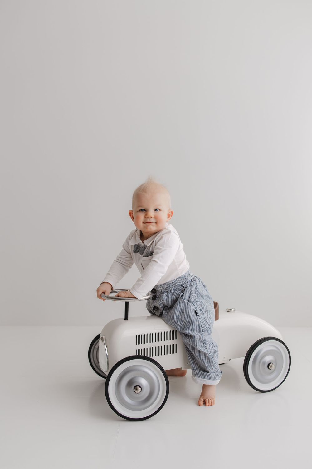 Smiling baby in white shirt and jeans riding a vintage white toy car in a bright studio setting.