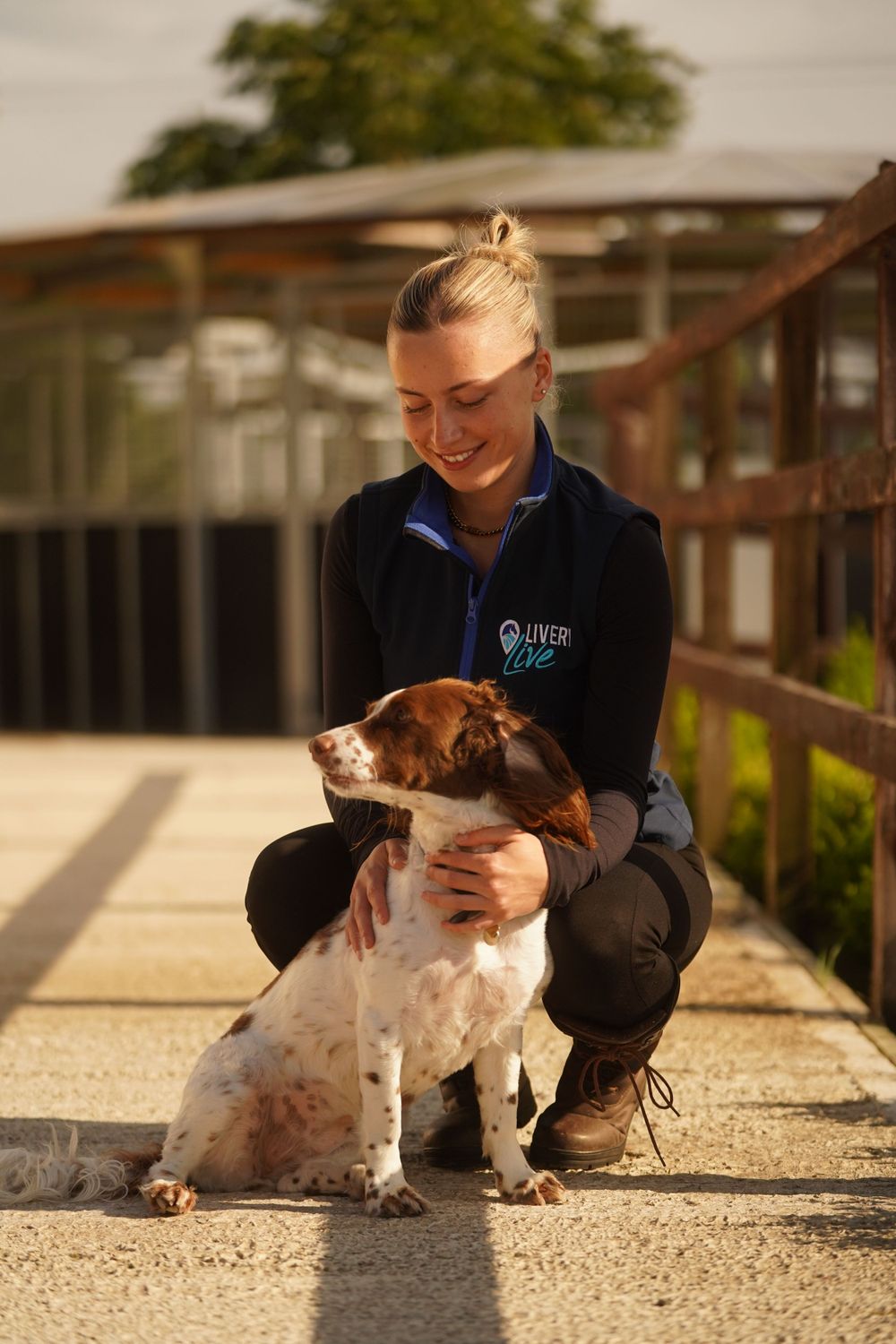 A dog lover in a blue uniform vest kneels down to pet a friendly spotted brown and white dog outdoors.