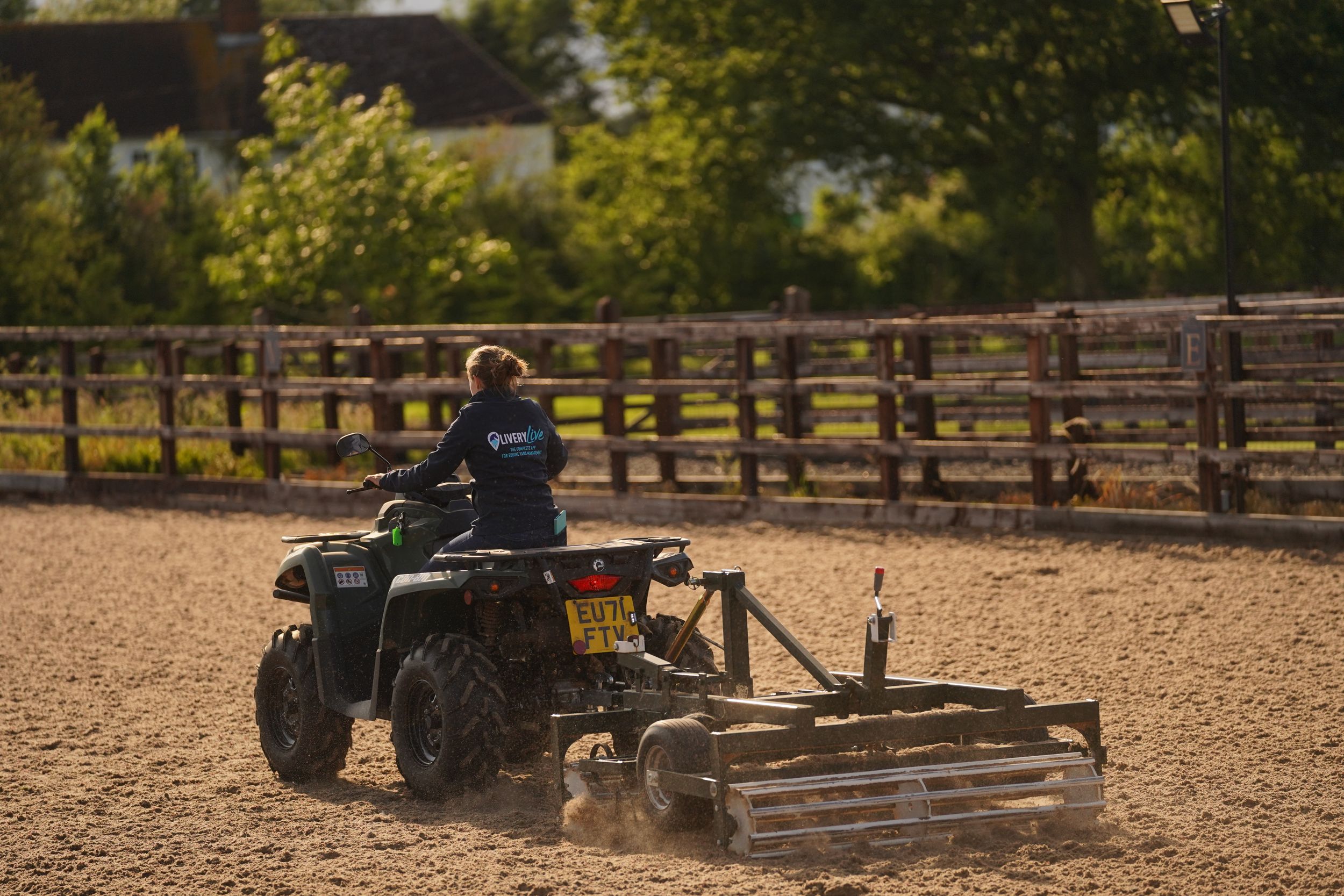 ATV rider operates a dirt leveling drag attachment in a fenced equestrian arena on a sunny day.