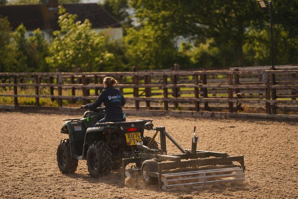 ATV rider operates a dirt leveling drag attachment in a fenced equestrian arena on a sunny day.
