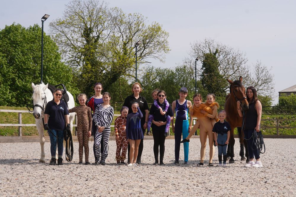 A group of people and horses stand together on a sandy riding arena surrounded by trees and fencing.