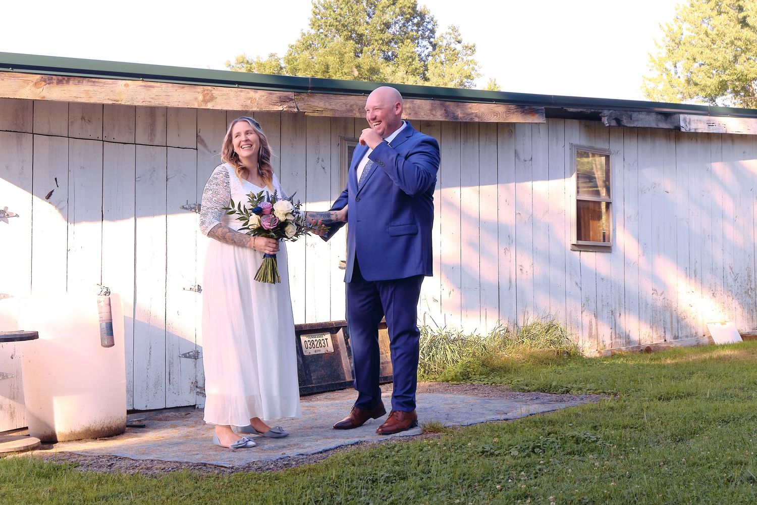 Two people standing together dressed in wedding attire outside a rustic white building on a sunny day.
