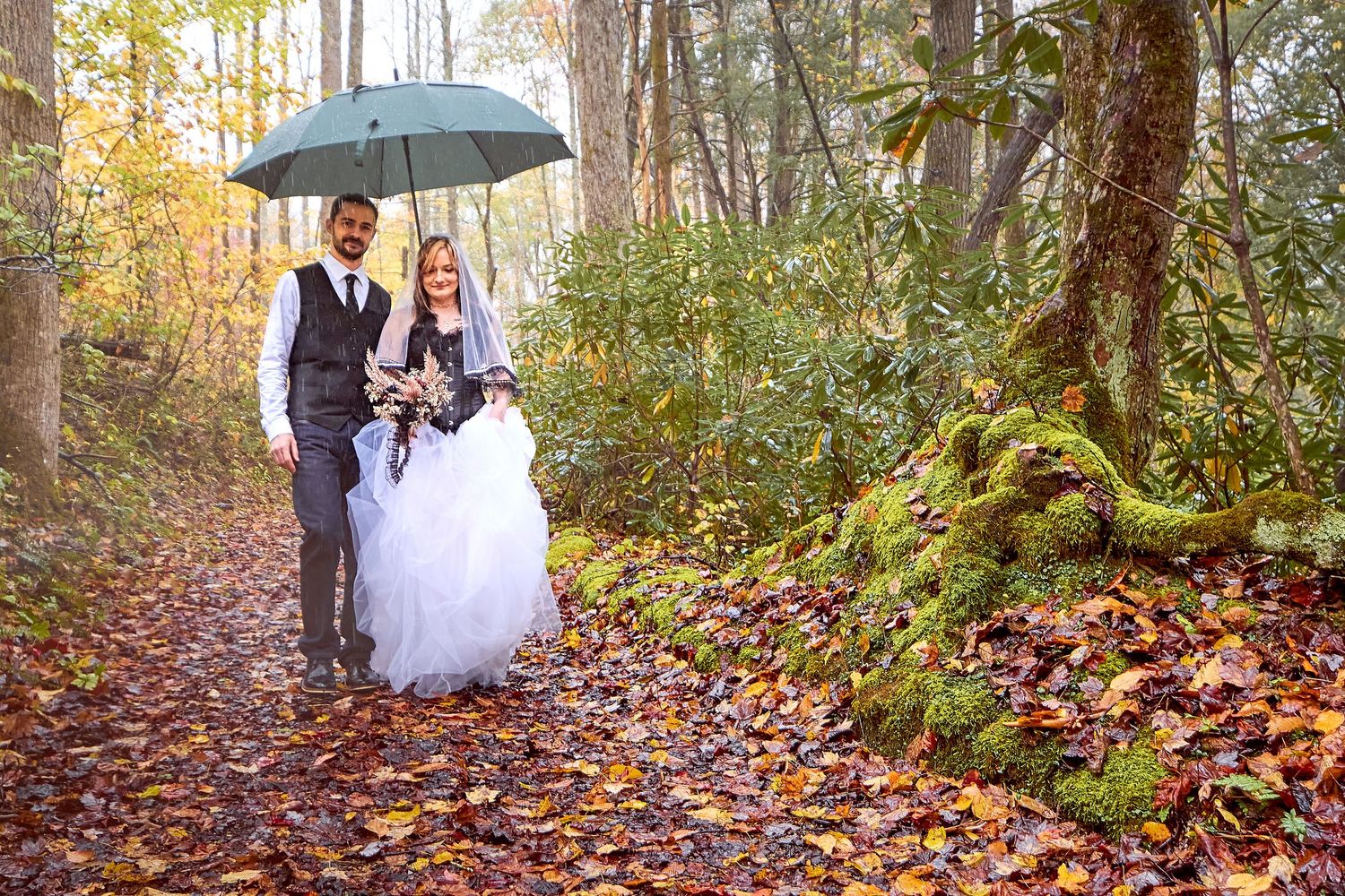 A couple in wedding attire walks along a rainy autumn forest path while holding a black umbrella.