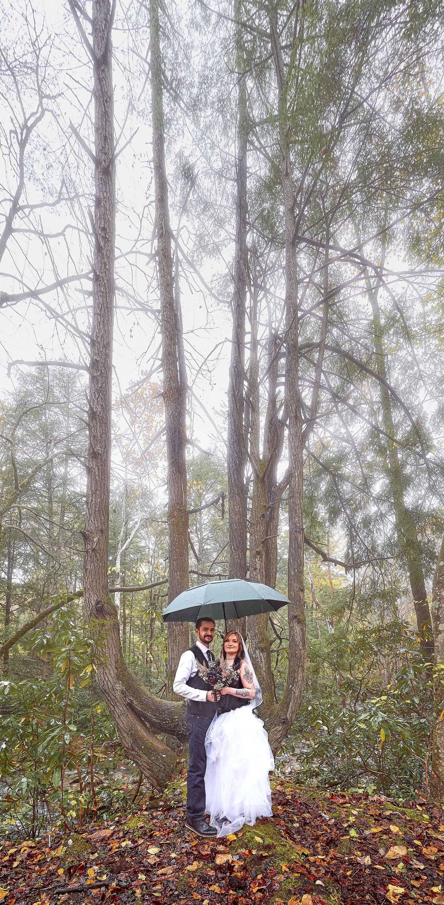 A couple shares a romantic moment in a misty forest with fallen autumn leaves and an umbrella.