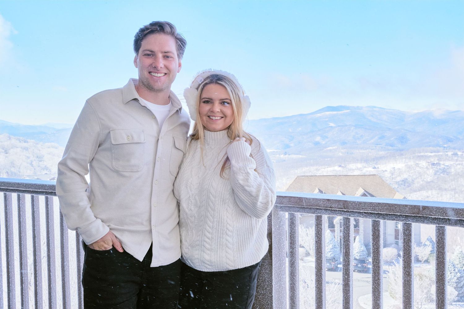 A couple stands together on a snowy mountain overlook with scenic winter views in the background.