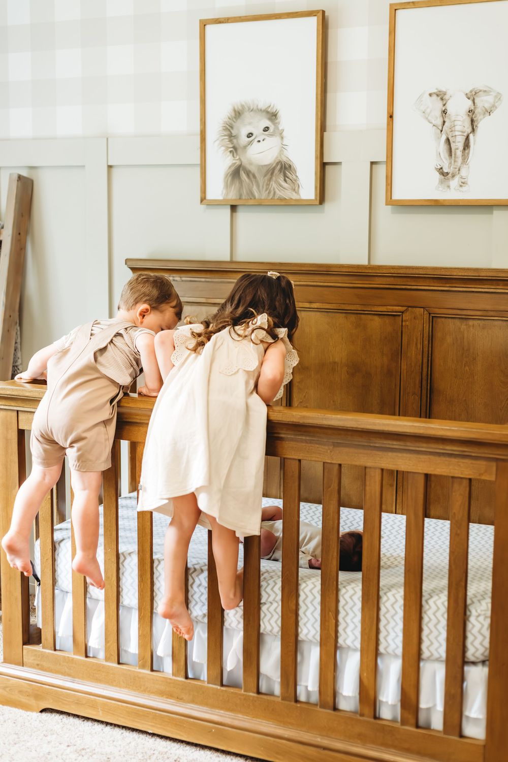 Two children in light colored dresses peeking into wooden crib beneath framed animal prints in New Haven, CT.