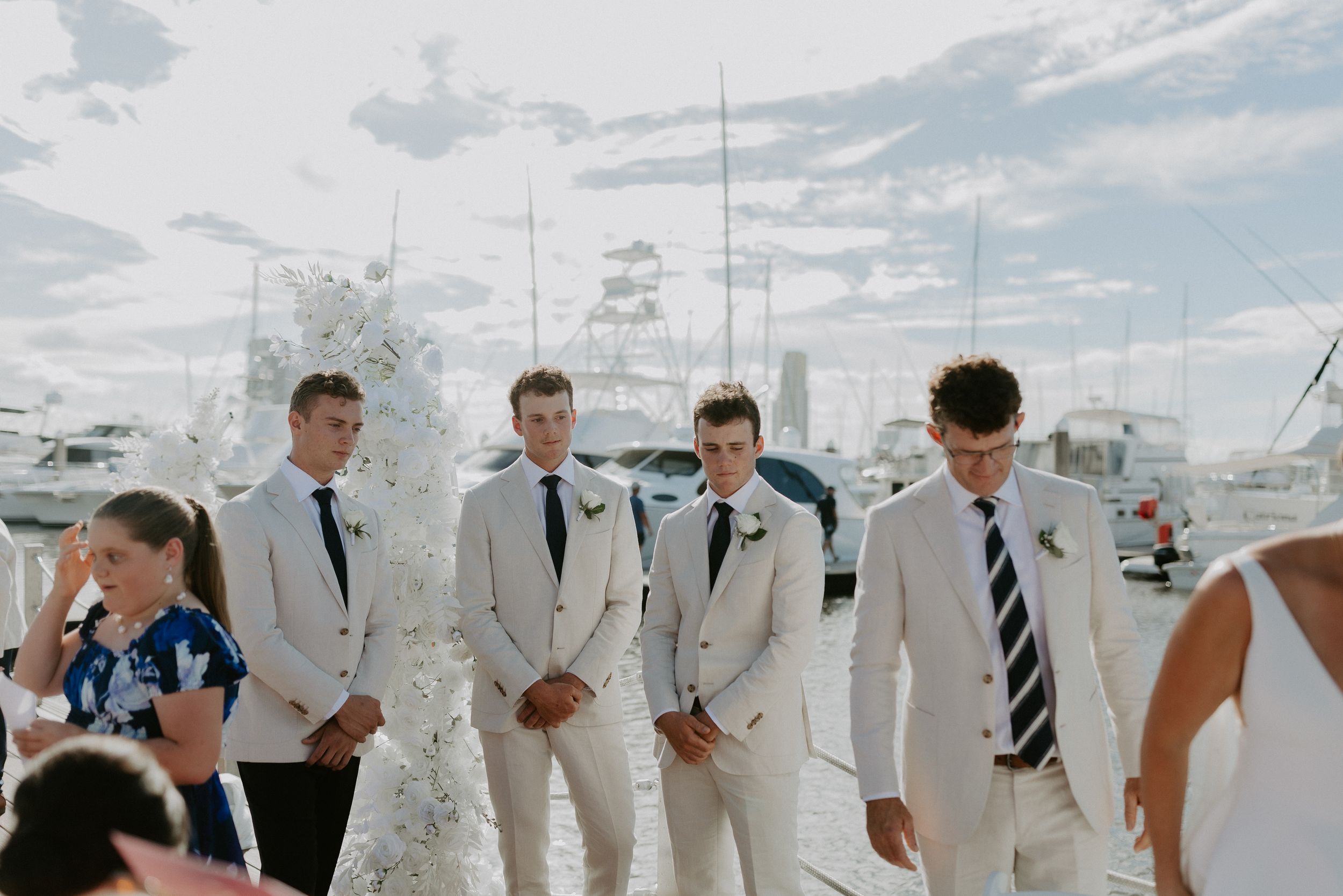 A group of four men in light-colored suits with dark ties are standing outdoors. They are near marina with boats in the background. The sky!
